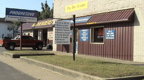 A white building wiht a brown metal siding facade with a small grass long along a street. Text on sign in front has text: - Auto and SR 22 - Motorcicles - Home and Farm - Mobile Homes - Workers Comp - Security Bonds - Life and Health - Medicare Supplements
							   Another sign has text: Progressive Realty and Insurance
							   Another sign has text Pay Day Loans
