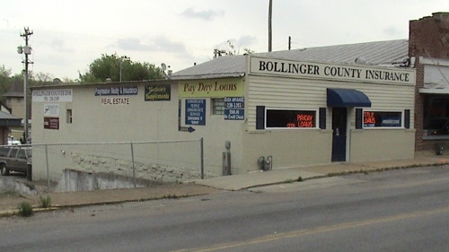 A white, one story building on a street. There are two windows and a single door facing the street and a sign that reads: Bollinger County Insurance