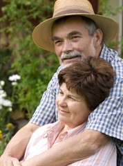 An older couple, man with beard and hat and woman leaning against him. A background of green foliage