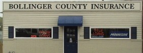 an off-white, one story building with 2 wide windows and blue door. The sign above has text: Bollinger County Insurance